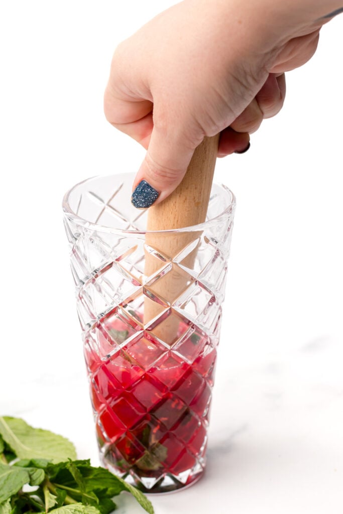 A hand uses a wooden muddler to mash red fruit and mint leaves in a patterned glass, with fresh mint visible beside the glass.