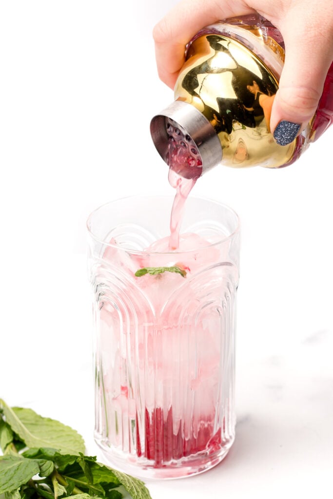 A hand pours pink liquid from a gold cocktail shaker into a glass filled with ice and mint leaves; fresh mint is on the surface nearby.