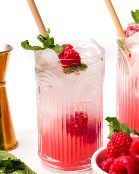 A glass of pink raspberry cocktail with ice, garnished with fresh mint and raspberries, with a bamboo straw. A golden jigger, mint leaves, and a bowl of raspberries are nearby.