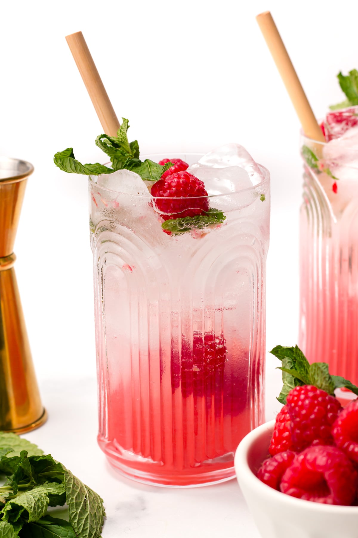 A glass of pink raspberry cocktail with ice, garnished with fresh mint and raspberries, with a bamboo straw. A golden jigger, mint leaves, and a bowl of raspberries are nearby.