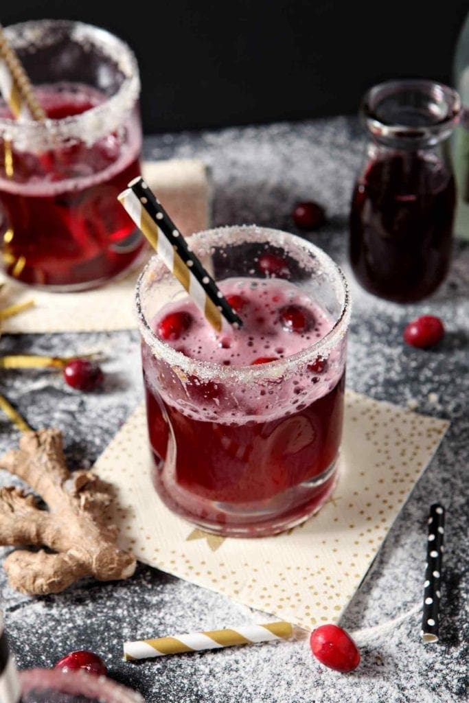 A glass of red cranberry drink with a sugar rim, black-and-yellow striped straw, surrounded by cranberries, ginger, and a small bottle of juice on a speckled surface.