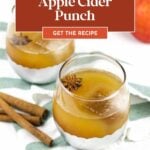 Two glasses of apple cider punch garnished with star anise, next to cinnamon sticks on a green striped cloth, with apples in the background.
