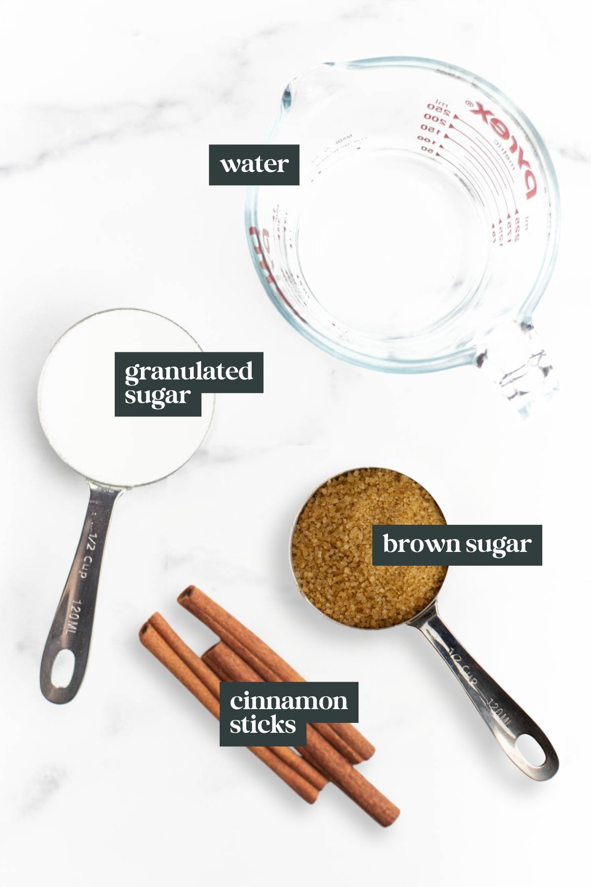 A measuring cup of water, a metal cup of granulated sugar, a metal cup of brown sugar, and three cinnamon sticks are arranged on a white surface.
