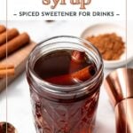 A glass jar filled with cinnamon syrup, garnished with a cinnamon stick, sits on a white surface next to a spoon, cinnamon sticks, and a bowl of ground cinnamon.