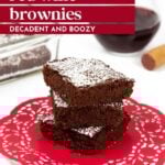 A stack of chocolate brownies dusted with powdered sugar sits on a red doily, with a glass of red wine and a baking dish in the background.