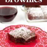 A piece of chocolate brownie with powdered sugar sits on a red doily, next to a glass of red wine and a dish of more brownies in the background.