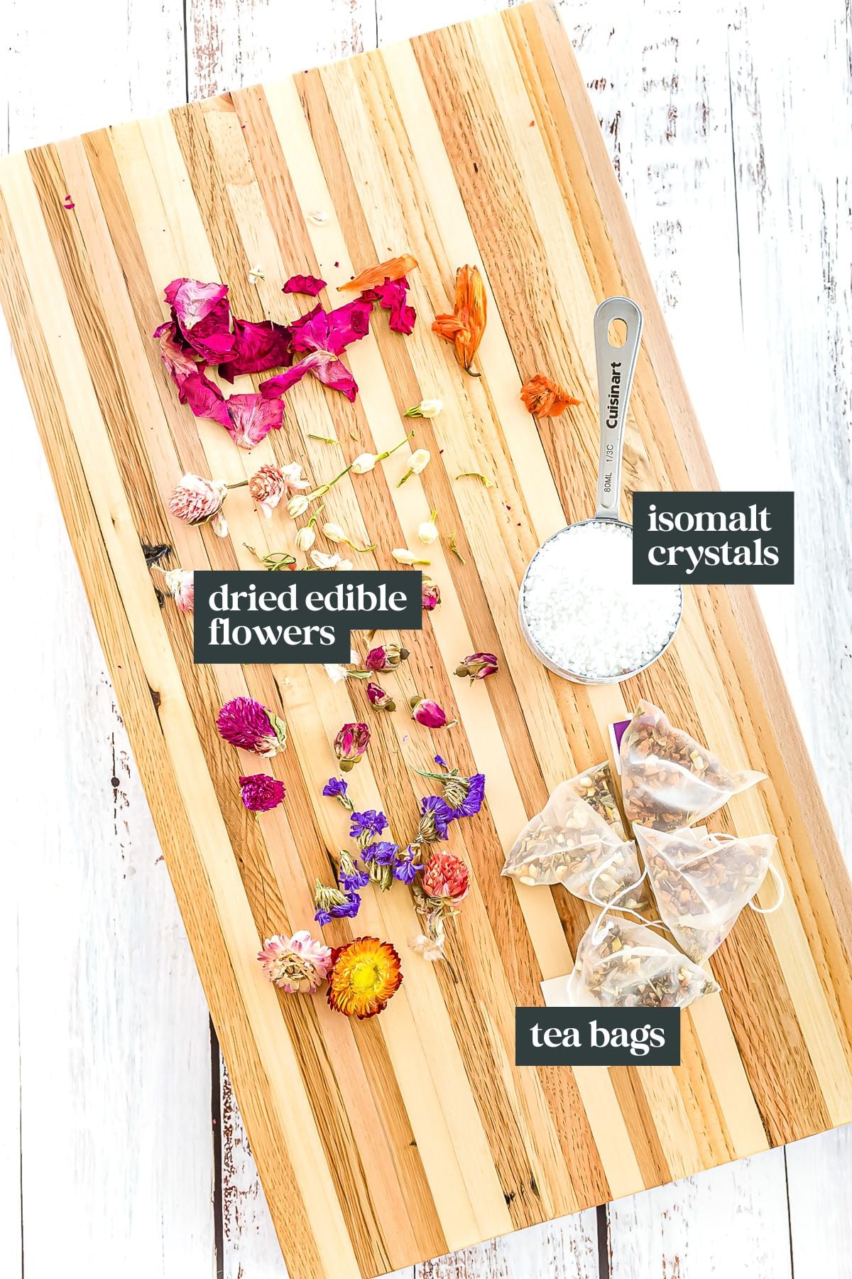 A wooden board displays dried edible flowers, a metal measuring cup of isomalt crystals, and several tea bags, each labeled for identification.