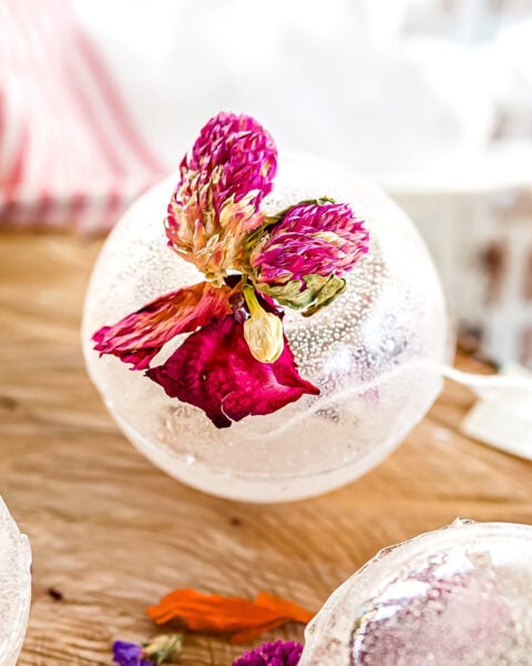 A clear ice sphere with a pink and purple dried flower frozen inside sits on a wooden surface.