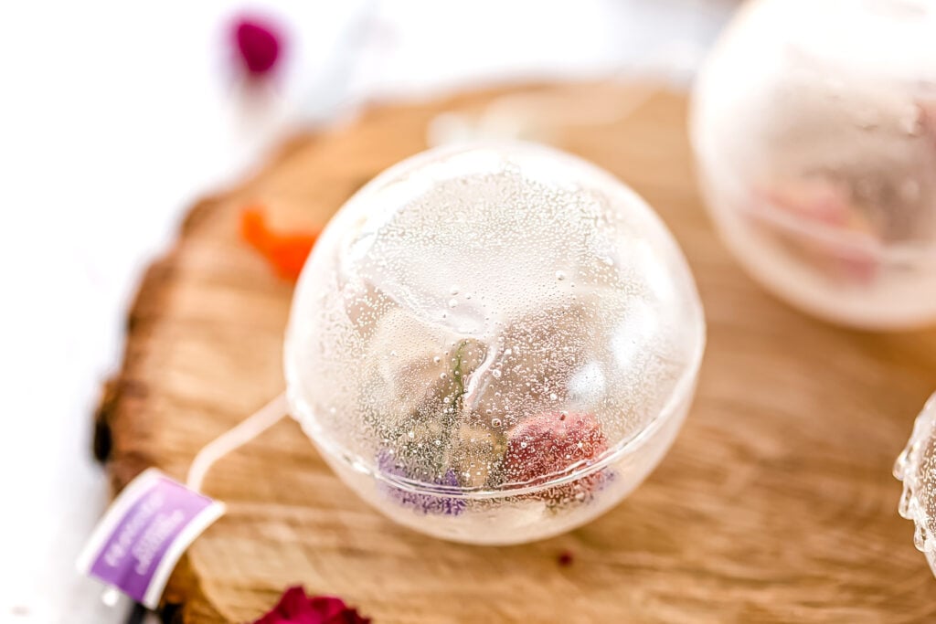 A clear spherical ornament with condensation on the surface sits on a wooden slab, containing small colorful dried flowers inside.
