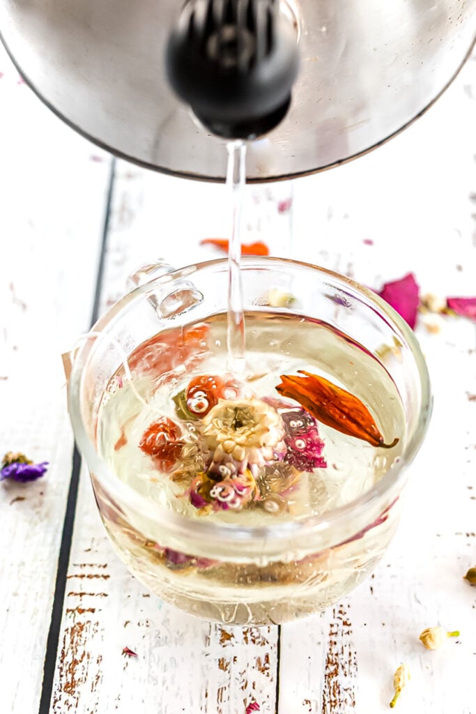 Hot water is being poured from a kettle into a glass cup containing a blooming floral tea on a white wooden surface.