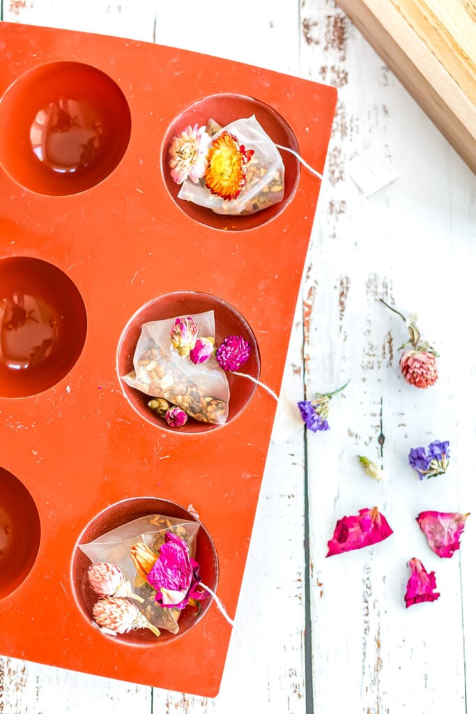 Red silicone mold with circular cavities contains sachets filled with dried flowers and herbs; loose petals and flowers scattered nearby on a white wooden surface.