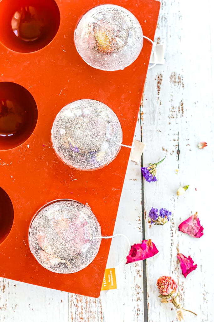 Three glittery tea bomb spheres rest in a red silicone mold on a white wooden surface, with scattered dried flower petals nearby.