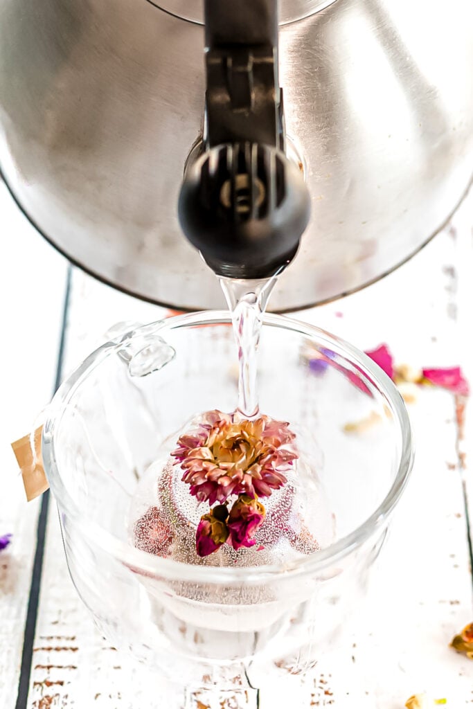 Hot water is being poured from a kettle into a glass cup containing a tea infuser with dried flowers, creating an experience similar to brewing tea bombs.