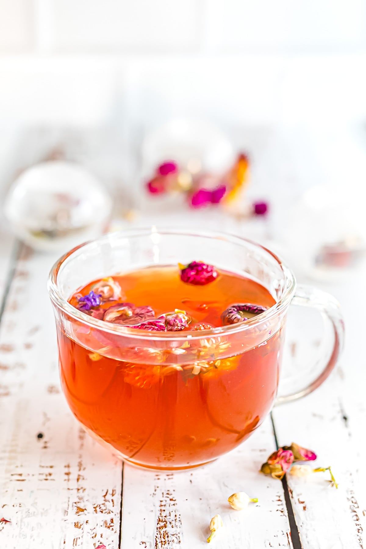 A clear glass cup filled with herbal tea, topped with dried flower buds, sits on a white rustic wooden surface.