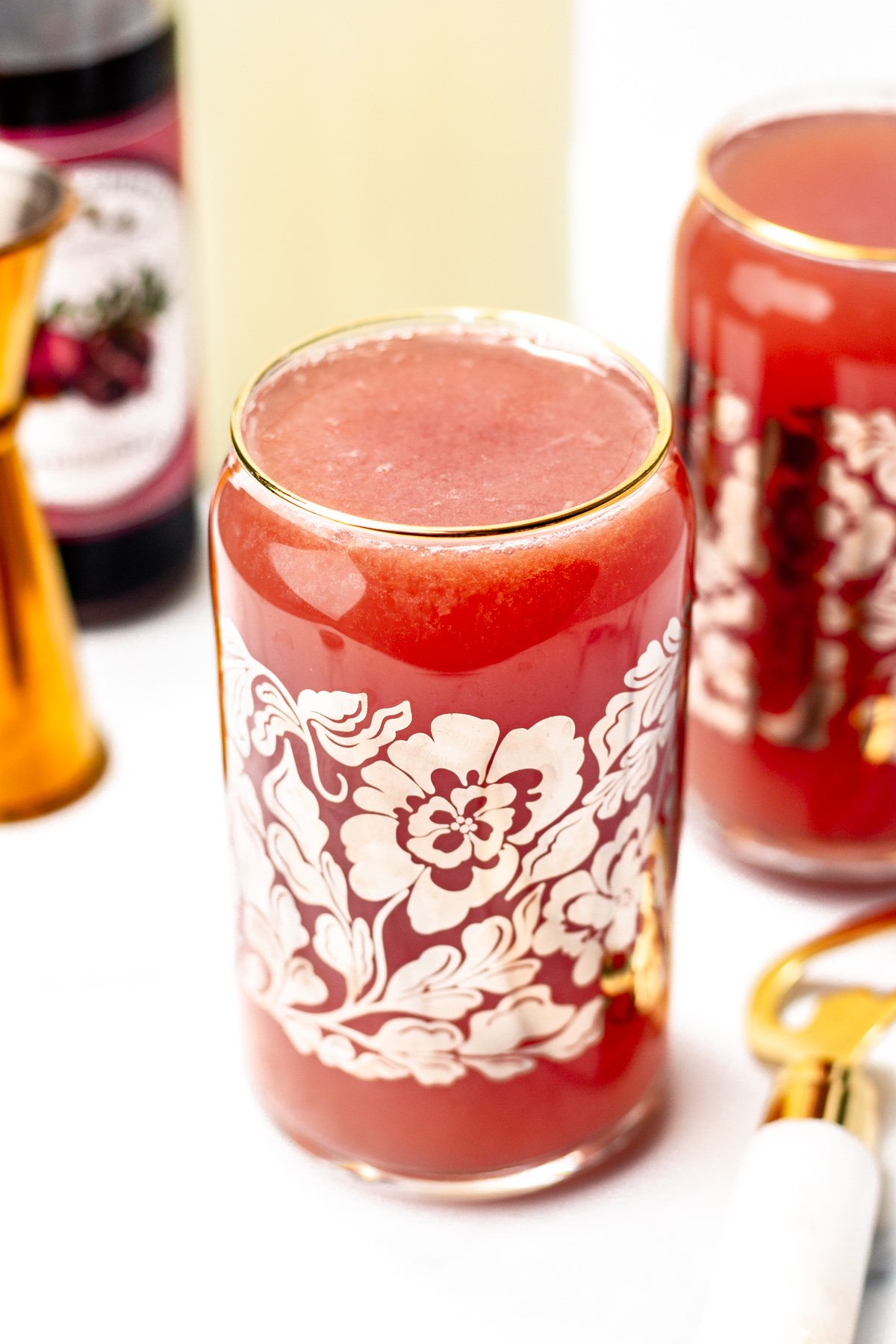 A glass with a floral design filled with a red beverage sits on a white surface, surrounded by bar tools and drink bottles.