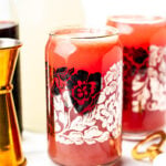 A glass with a red floral design filled with a red beverage sits on a white surface, surrounded by a jigger, bottle, and another glass in the background.