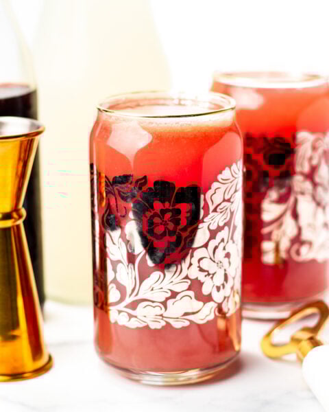 A glass with a red floral design filled with a red beverage sits on a white surface, surrounded by a jigger, bottle, and another glass in the background.