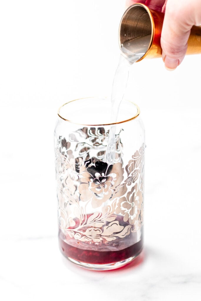 A hand pours clear liquid from a jigger into a glass with floral patterns, containing a small amount of red liquid at the bottom.