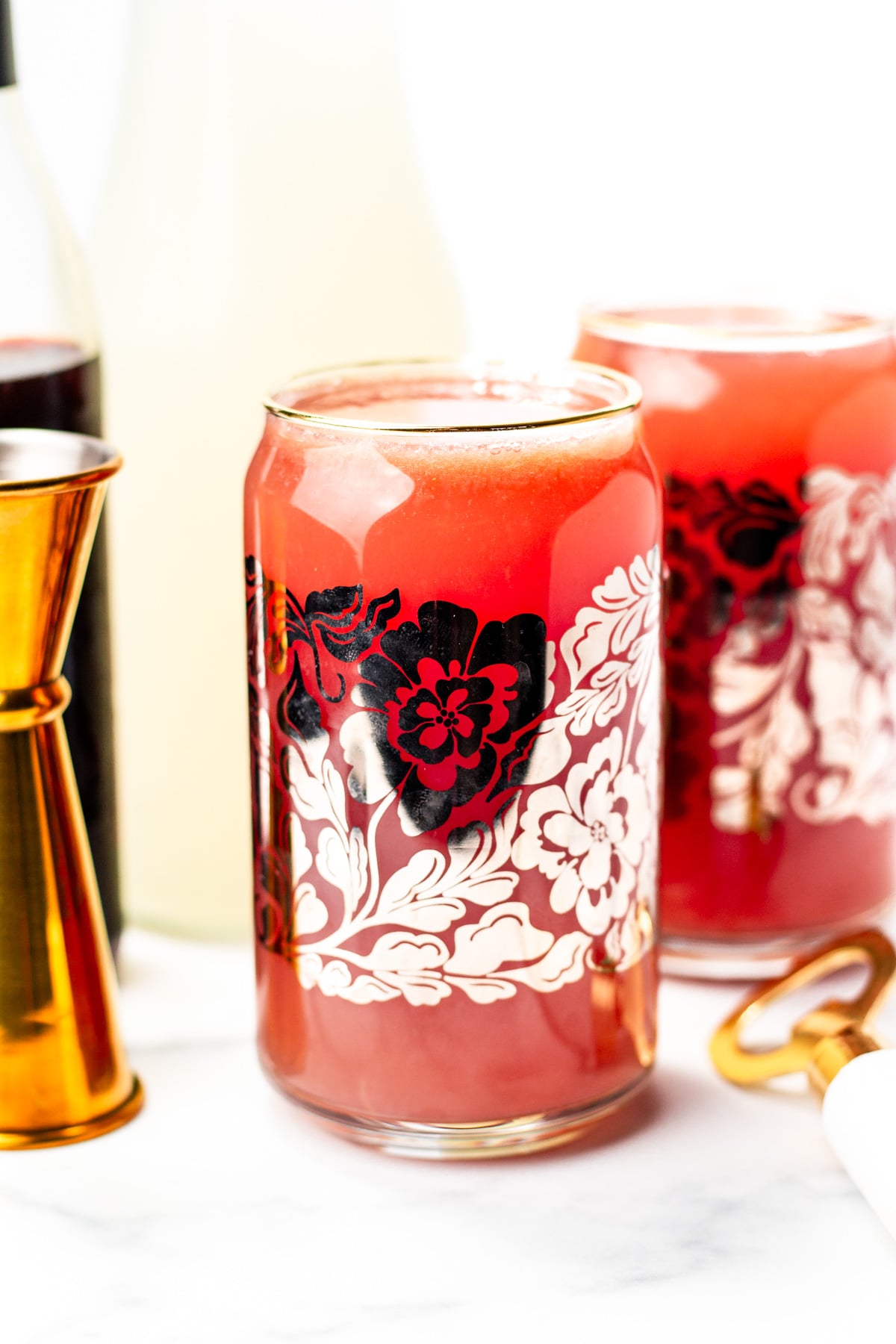A glass with a red floral design filled with a red beverage sits on a white surface, surrounded by a jigger, bottle, and another glass in the background.