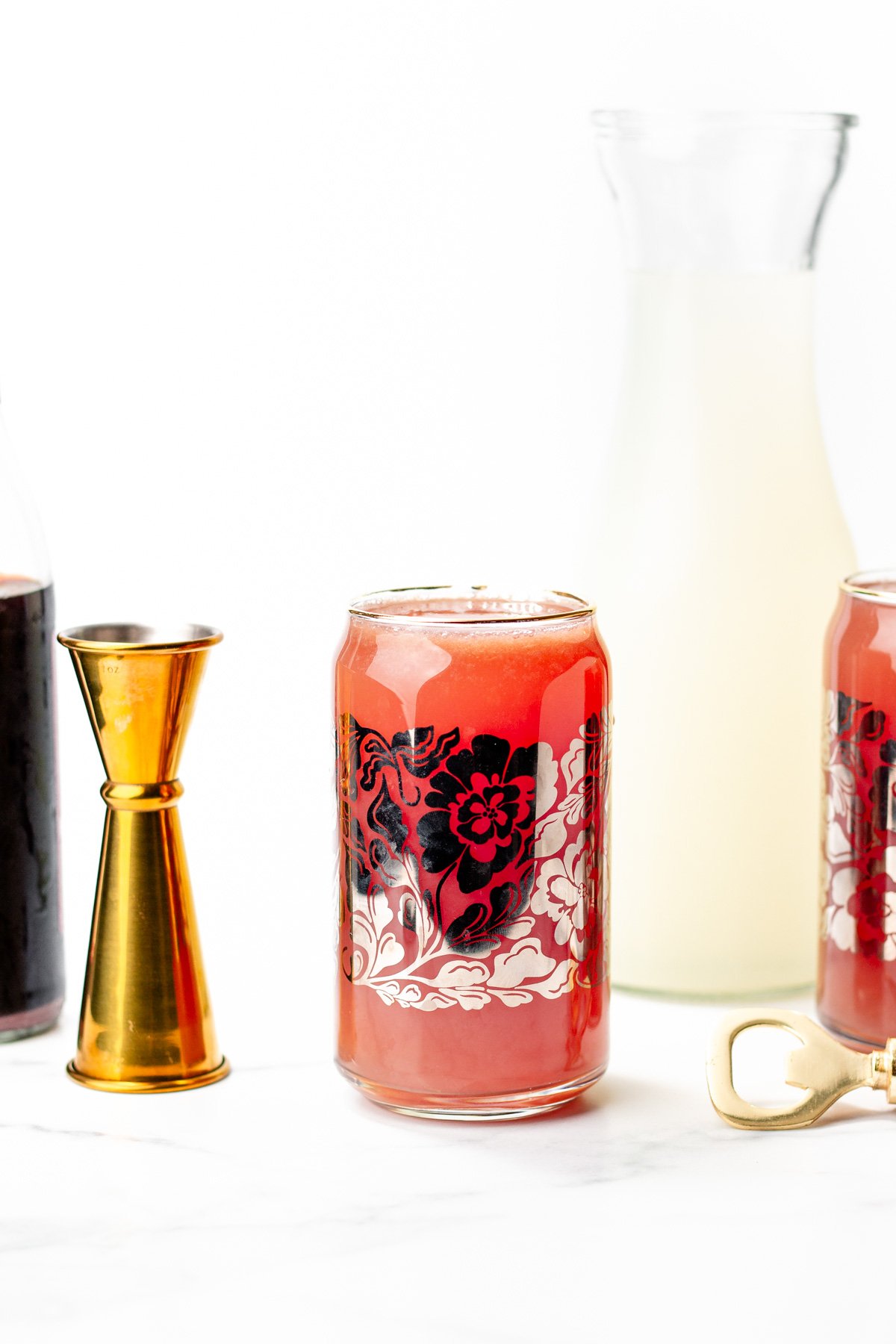 A floral-patterned glass filled with a red drink is on a white surface alongside a gold jigger, a carafe of pale liquid, another glass, and a bottle opener.