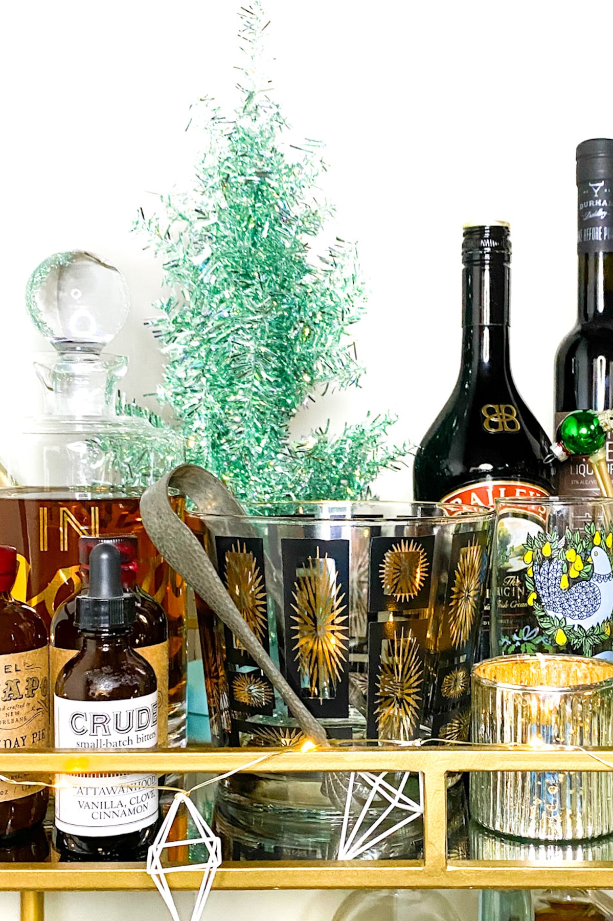 A festive bar cart with assorted liquor bottles, a glass with gold starburst designs, cocktail bitters, and a small green decorative tree.