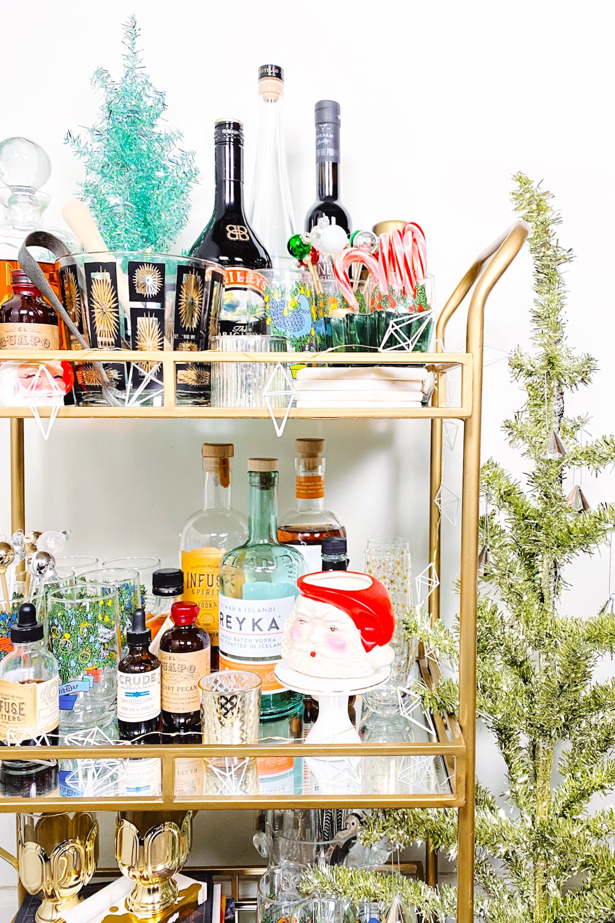 A gold bar cart decorated for the holidays with various liquor bottles, cocktail glasses, a Santa mug, candy canes, small Christmas trees, and a festive stocking&mdash;a charming addition to your home bar this season.