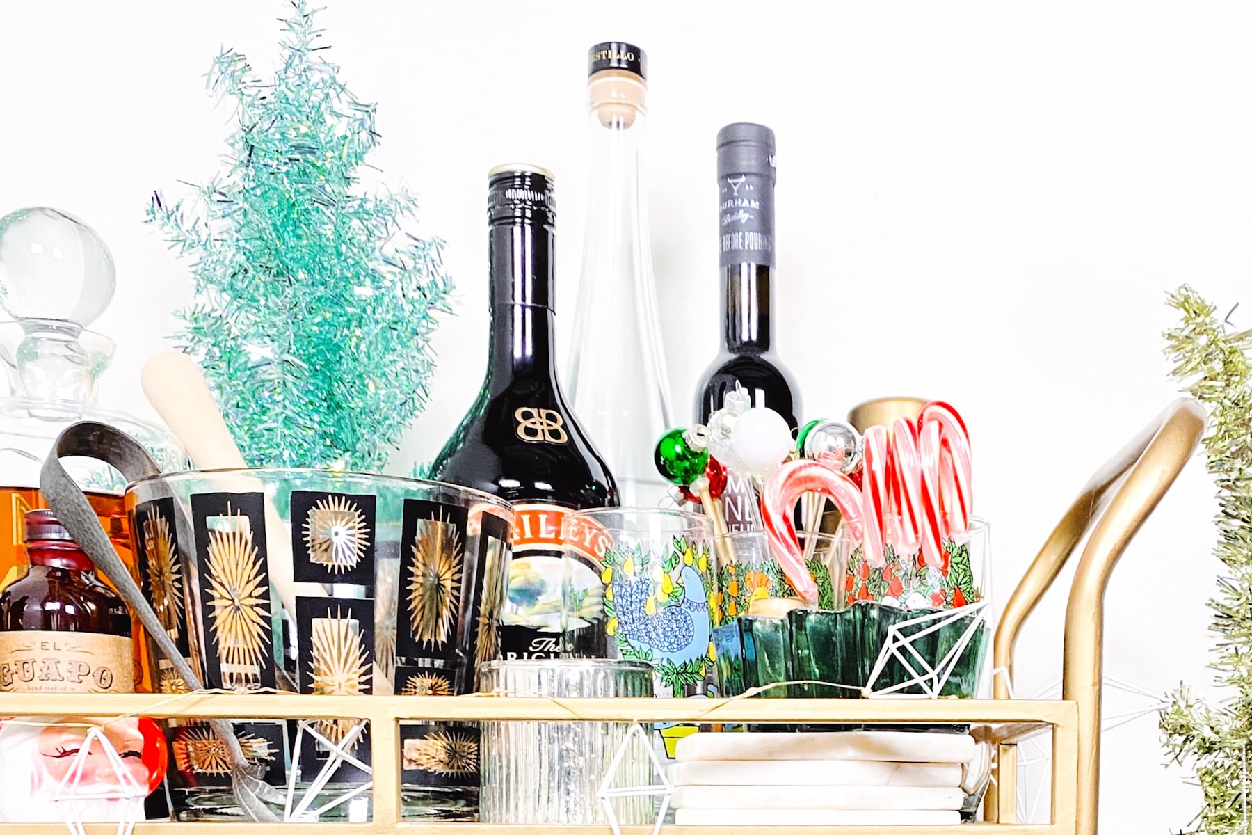 A gold bar cart with assorted liquor bottles, glasses, candy canes, and festive decorations arranged on top.