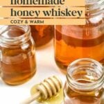 Jars filled with golden homemade honey whiskey sit on a white surface, with a wooden honey dipper and some honey in the foreground.
