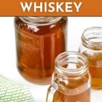 A large glass jar and two small mason jars filled with honey whiskey are displayed on a white surface next to a green patterned cloth. Text reads “Homemade Honey Whiskey. Get the recipe. feastandwest.com.”.