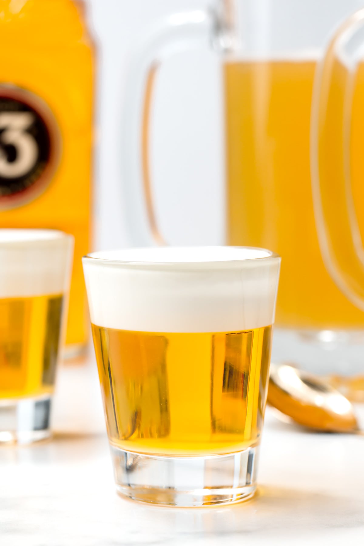 A glass filled with a golden yellow beverage topped with a layer of white foam, with similar drinks and a pitcher in the background.