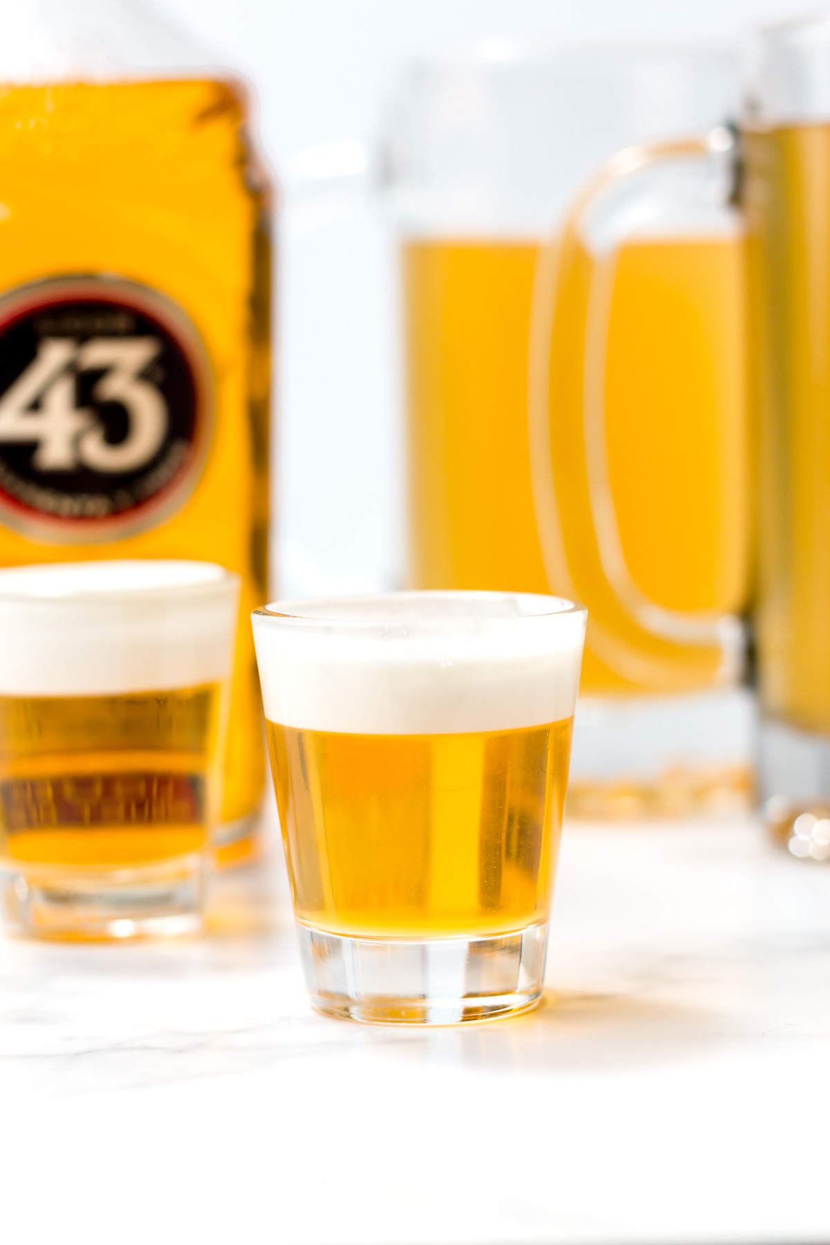 A close-up of a shot glass with golden liquid and a creamy white foam on top, with bottles and mugs of similar drinks in the background.
