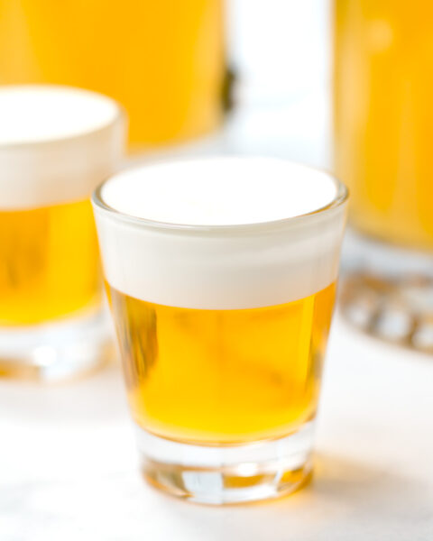 A clear shot glass filled with a golden yellow liquid topped with a layer of white foam, placed on a white surface.