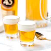 Two layered shots with a yellow liquid base and a creamy white top sit on a white surface, surrounded by a bottle, a glass pitcher, a spoon, and a sugar container.