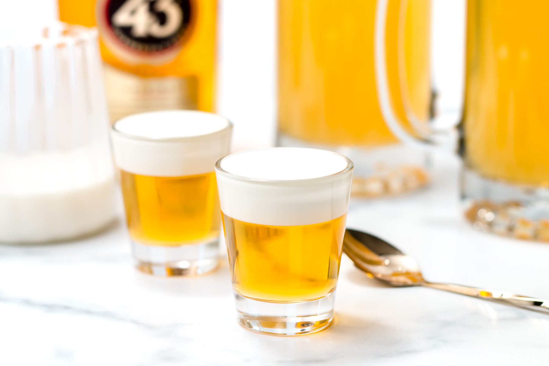 Two layered shots with a yellow liquid base and a creamy white top sit on a white surface, surrounded by a bottle, a glass pitcher, a spoon, and a sugar container.