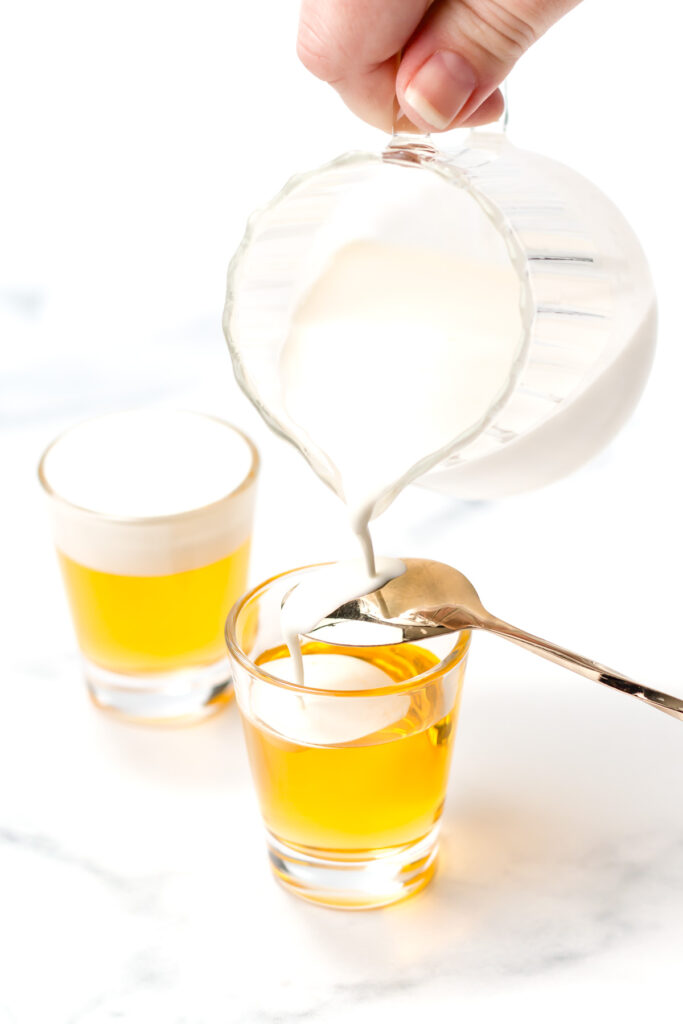 A hand pours cream from a glass pitcher over the back of a spoon into a shot glass filled with a golden liquid, next to a similar prepared drink.