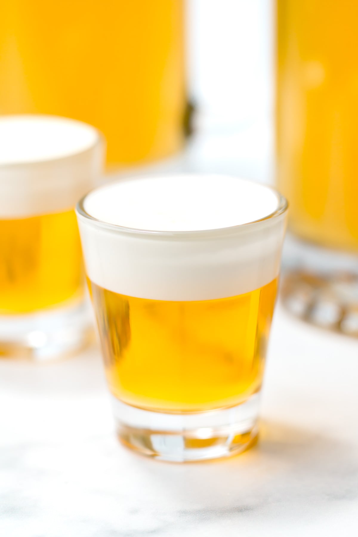 A clear shot glass filled with a golden yellow liquid topped with a layer of white foam, placed on a white surface.
