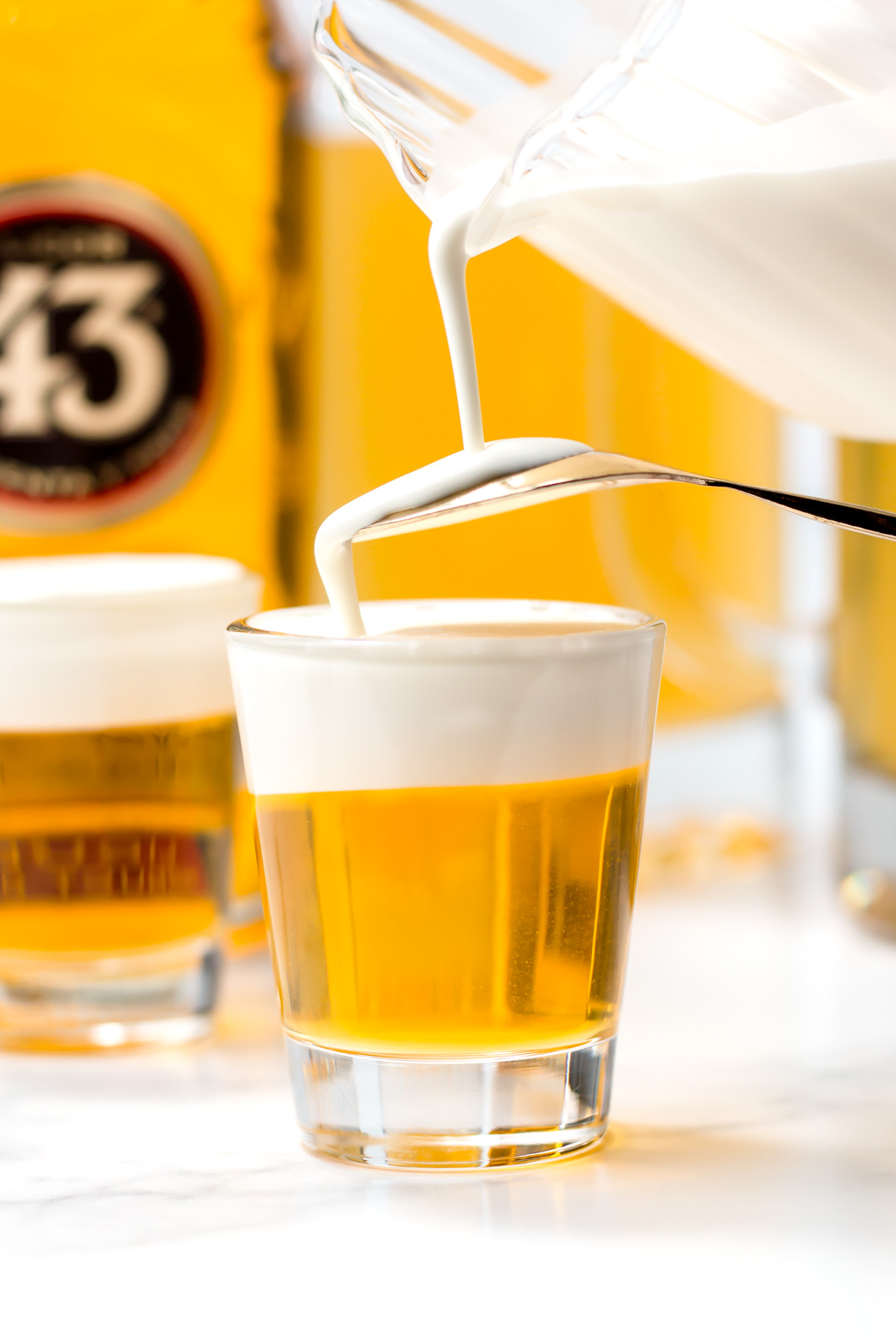 A close-up of a clear shot glass filled with yellow liqueur as white cream is being poured on top, creating a layered drink. Bottles are visible in the blurred background.