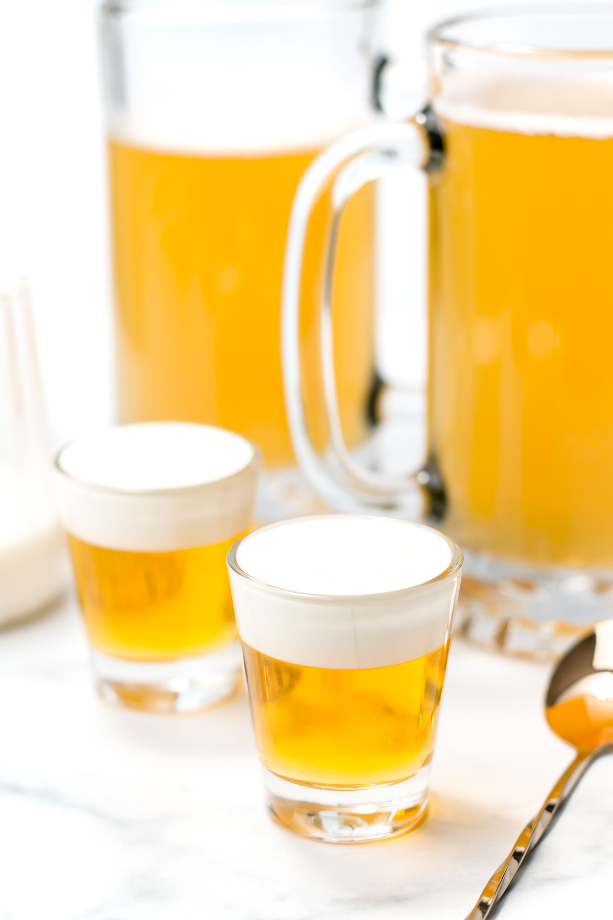 Two shot glasses with a yellow liquid topped with white foam are placed in front of large beer mugs filled with a similar drink on a white surface.