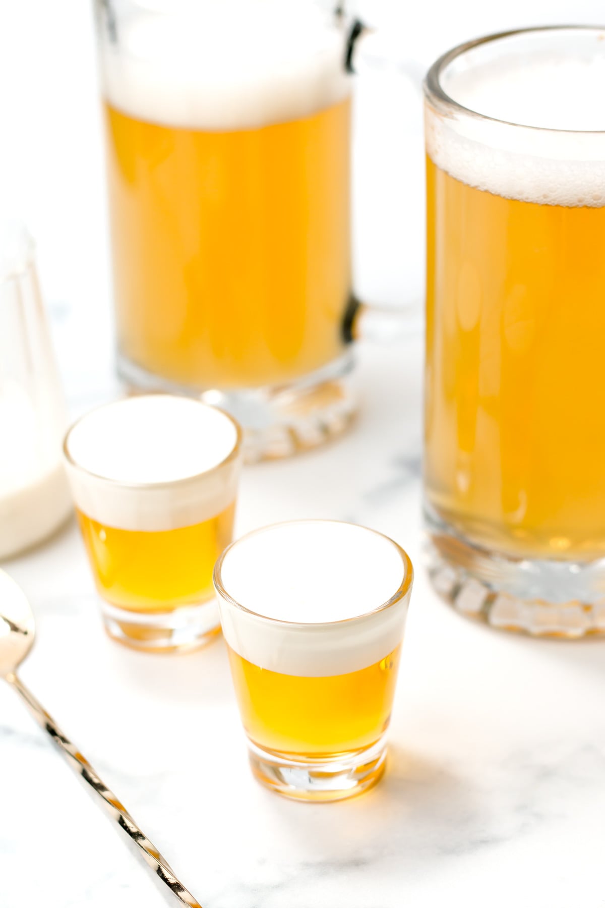 Two shot glasses with layered yellow and white drinks are in the foreground, with two glass mugs of light beer in the background on a white surface.