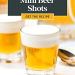 Close-up of two mini beer shots with creamy foam on top, a spoon, and a partial drink in the background. Text overlay reads: "Mini Beer Shots. Get the Recipe.