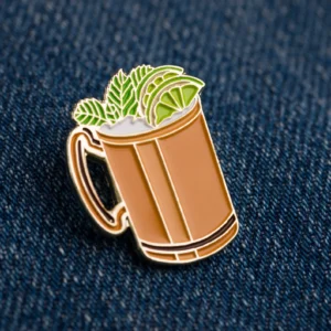 A metal pin shaped like a copper mug with lime wedges, mint leaves, and ice, placed on dark blue denim fabric.
