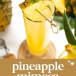 A glass of pineapple mimosa garnished with a pineapple slice and leaf, set on a wooden coaster with fresh pineapple in the background. Text overlay reads "pineapple mimosa, tropical & tangy.