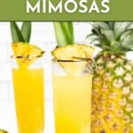 Two tall glasses of yellow pineapple mimosa garnished with pineapple slices and leaves, with a whole pineapple in the background. Text reads "Pineapple Mimosas - Get the Recipe.