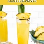 A tall glass of pineapple mimosa garnished with a pineapple slice and leaf sits on a coaster; another glass and pineapple pieces are in the background.