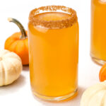A glass of orange-colored drink with a brown sugared rim, surrounded by small orange and white pumpkins on a white surface.
