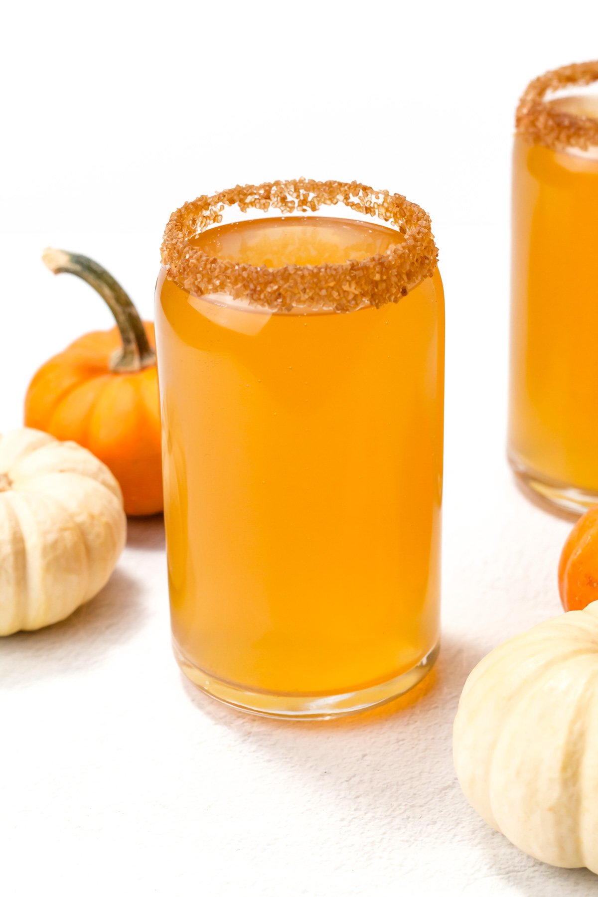 A glass of orange-colored drink with a brown sugared rim, surrounded by small orange and white pumpkins on a white surface.