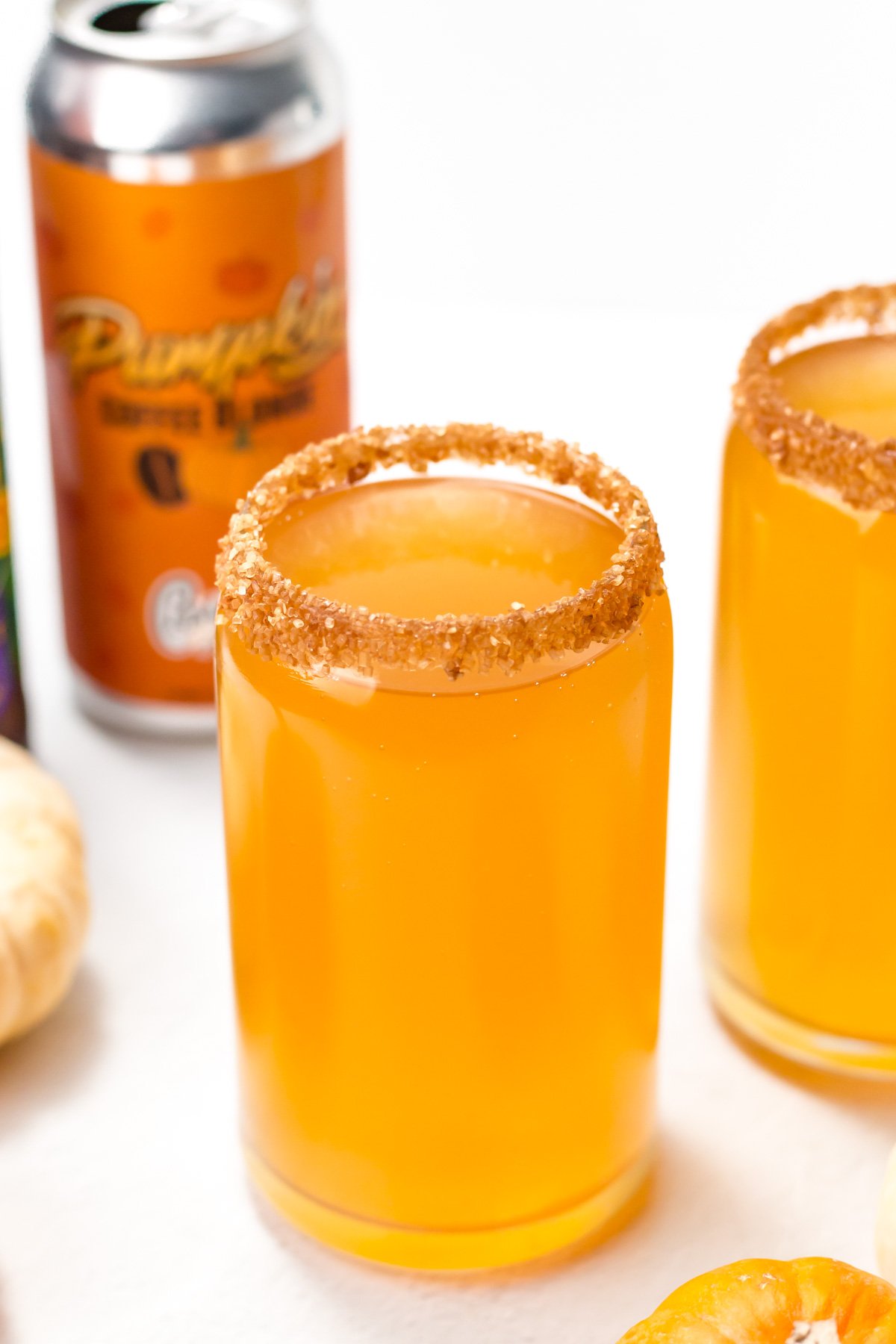 A glass of orange pumpkin beer with a spiced sugar rim, with a can labeled "Pumpkin" and small pumpkins in the background.