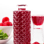 A glass decanter filled with red liquid sits beside a glass, raspberries, and mint leaves on a white surface.