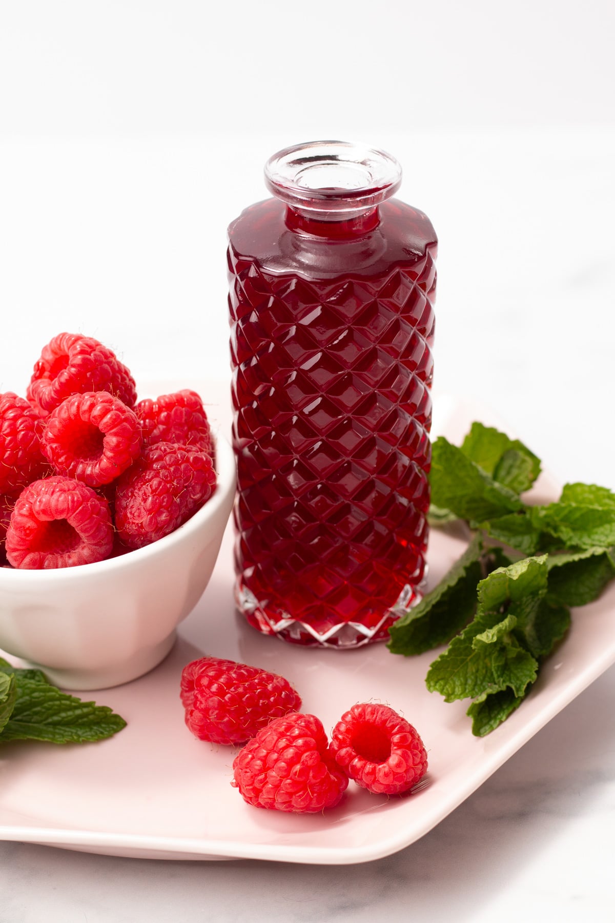 A glass bottle of raspberry syrup stands next to a bowl of fresh raspberries and mint leaves on a pink plate.