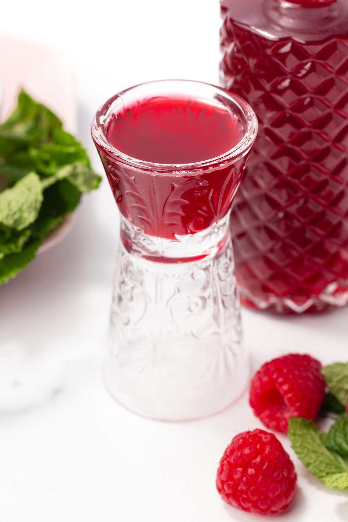 A clear glass filled with red liquid sits on a white surface next to fresh raspberries, mint leaves, and a patterned bottle containing more red liquid.
